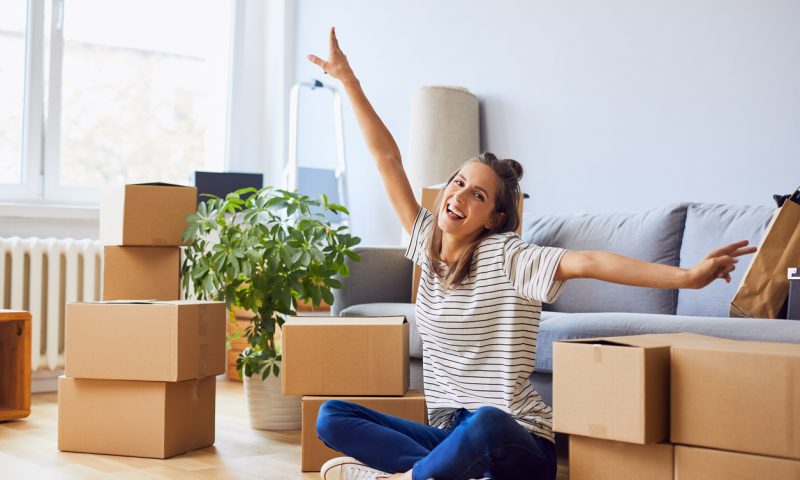 Young woman sitting in new apartment and raising arms in joy after moving in