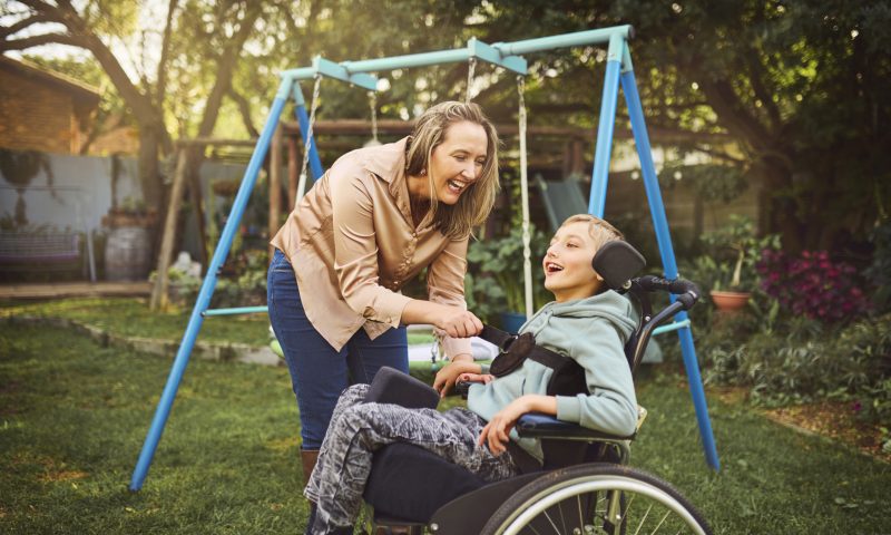 Woman wearing casual attire, leaning forward smiling cheerfully, calmly removing strap from happy young special son sitting in wheelchair, enjoying morning air and sun in garden